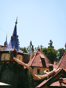 A whimsical building with tall, pointed roofs covered in blue and red tiles, resembling a fairytale castle. Long, golden hair drapes from one of the windows, hinting at a storybook theme. Surrounding the structure are trees under a clear blue sky.