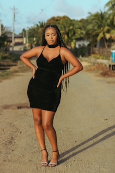 A woman in a black dress with long braids stands confidently on a dirt road. The background includes palm trees and a few houses, suggesting a tropical location. The setting sun casts a warm glow, enhancing the natural hues.