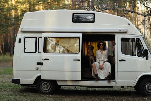 A person wearing sunglasses and a white dress sits relaxed in the doorway of a white camper van, with a forest setting in the background. The van is parked on grass surrounded by trees, and the warm light suggests a peaceful atmosphere.