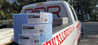 Two rolls of Fibafuse Max drywall tape are placed on the open tailgate of a white pickup truck. The truck has a logo with red text on the side. In the background, there are trees and another vehicle is visible.
