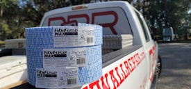 Two rolls of Fibafuse Max drywall tape are placed on the open tailgate of a white pickup truck. The truck has a logo with red text on the side. In the background, there are trees and another vehicle is visible.