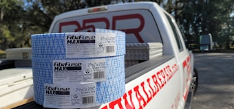 Two rolls of Fibafuse Max drywall tape are placed on the open tailgate of a white pickup truck. The truck has a logo with red text on the side. In the background, there are trees and another vehicle is visible.