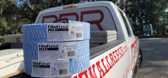 Two rolls of Fibafuse Max drywall tape are placed on the open tailgate of a white pickup truck. The truck has a logo with red text on the side. In the background, there are trees and another vehicle is visible.
