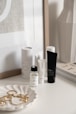 A collection of skincare and beauty products is displayed on a white surface. There are bottles and tubes of various sizes, with labels visible on some of them. A decorative white dish holds a selection of gold jewelry pieces, including earrings and perhaps a necklace. The background includes a framed print and the corner of a magazine stack.