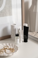 A collection of skincare and beauty products is displayed on a white surface. There are bottles and tubes of various sizes, with labels visible on some of them. A decorative white dish holds a selection of gold jewelry pieces, including earrings and perhaps a necklace. The background includes a framed print and the corner of a magazine stack.