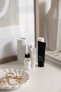A collection of skincare and beauty products is displayed on a white surface. There are bottles and tubes of various sizes, with labels visible on some of them. A decorative white dish holds a selection of gold jewelry pieces, including earrings and perhaps a necklace. The background includes a framed print and the corner of a magazine stack.