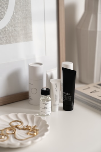 A collection of skincare and beauty products is displayed on a white surface. There are bottles and tubes of various sizes, with labels visible on some of them. A decorative white dish holds a selection of gold jewelry pieces, including earrings and perhaps a necklace. The background includes a framed print and the corner of a magazine stack.