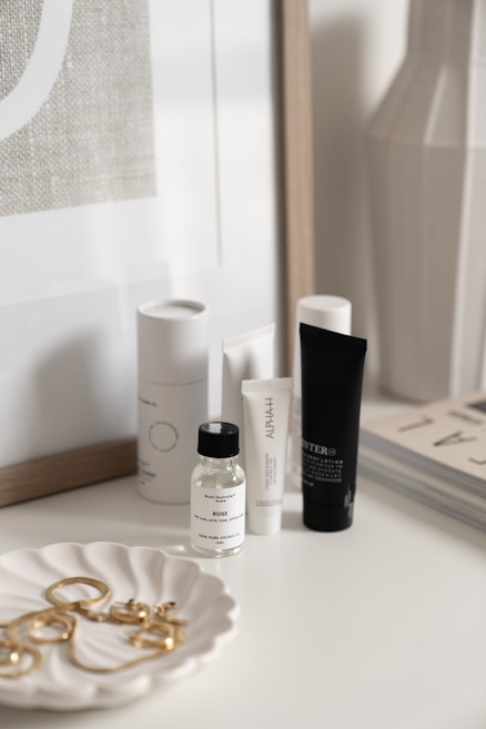 A collection of skincare and beauty products is displayed on a white surface. There are bottles and tubes of various sizes, with labels visible on some of them. A decorative white dish holds a selection of gold jewelry pieces, including earrings and perhaps a necklace. The background includes a framed print and the corner of a magazine stack.