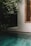 An outdoor scene featuring a tranquil swimming pool bordered by lush greenery and the side of a building with a wooden-framed window and shutters. The area is shaded, creating a serene and private atmosphere.