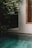 An outdoor scene featuring a tranquil swimming pool bordered by lush greenery and the side of a building with a wooden-framed window and shutters. The area is shaded, creating a serene and private atmosphere.