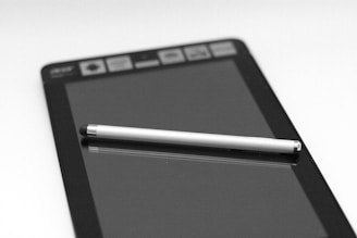 A tablet is placed on a flat surface with a stylus resting diagonally across its screen. The device displays a set of icons along the top edge. The black and white composition provides a sleek, modern feel.