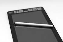 A tablet is placed on a flat surface with a stylus resting diagonally across its screen. The device displays a set of icons along the top edge. The black and white composition provides a sleek, modern feel.