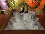 A intricately designed white wedding cake adorned with floral patterns and decorative swirls. A black silhouette cake topper depicting a couple is placed on the top tier. The backdrop features elaborate golden decorations with colorful flower arrangements.