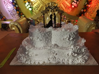 A intricately designed white wedding cake adorned with floral patterns and decorative swirls. A black silhouette cake topper depicting a couple is placed on the top tier. The backdrop features elaborate golden decorations with colorful flower arrangements.