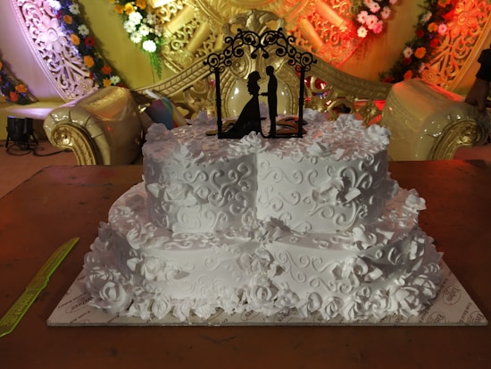 A intricately designed white wedding cake adorned with floral patterns and decorative swirls. A black silhouette cake topper depicting a couple is placed on the top tier. The backdrop features elaborate golden decorations with colorful flower arrangements.