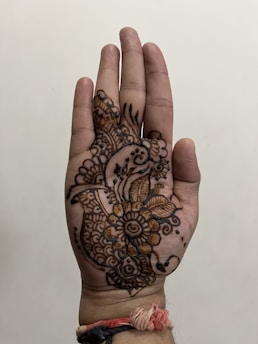 A hand is decorated with intricate mehndi designs, featuring floral patterns and paisley motifs. The henna art covers the palm and extends towards the fingers. A woven thread bracelet with red, white, and black strands is tied around the wrist.