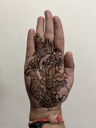 A hand is decorated with intricate mehndi designs, featuring floral patterns and paisley motifs. The henna art covers the palm and extends towards the fingers. A woven thread bracelet with red, white, and black strands is tied around the wrist.