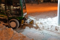A snow plow is parked on a street, clearing snow. The machinery is green and black with a large plow at the front pushing through the snow. The ground is covered with a thick layer of snow, and the surrounding area is dimly lit by orange street lights, creating a warm contrast against the cold environment.