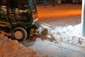 A snow plow is parked on a street, clearing snow. The machinery is green and black with a large plow at the front pushing through the snow. The ground is covered with a thick layer of snow, and the surrounding area is dimly lit by orange street lights, creating a warm contrast against the cold environment.