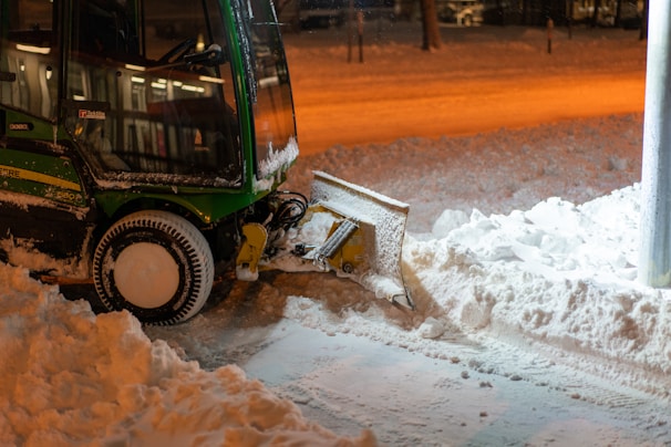 A snow plow is parked on a street, clearing snow. The machinery is green and black with a large plow at the front pushing through the snow. The ground is covered with a thick layer of snow, and the surrounding area is dimly lit by orange street lights, creating a warm contrast against the cold environment.