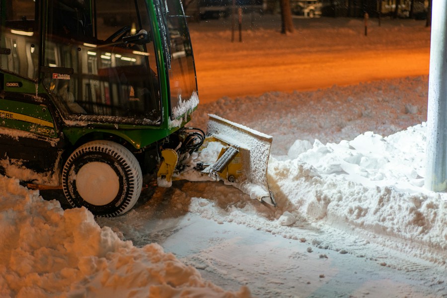 A snow plow is parked on a street, clearing snow. The machinery is green and black with a large plow at the front pushing through the snow. The ground is covered with a thick layer of snow, and the surrounding area is dimly lit by orange street lights, creating a warm contrast against the cold environment.