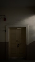 A closed wooden door with multiple panels is situated in a dimly lit narrow hallway. There is a black padlock securing the door handles. The wall surrounding the door is plain, with parts of it partially illuminated by soft light.