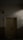 A closed wooden door with multiple panels is situated in a dimly lit narrow hallway. There is a black padlock securing the door handles. The wall surrounding the door is plain, with parts of it partially illuminated by soft light.