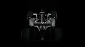 A sleek and aerodynamic Formula 1 car viewed from the front against a dark background. The car's contours and design elements are accentuated by the lighting, highlighting its sophisticated features.