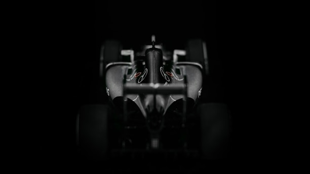 A sleek and aerodynamic Formula 1 car viewed from the front against a dark background. The car's contours and design elements are accentuated by the lighting, highlighting its sophisticated features.