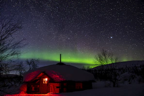 A peaceful snowy cabin illuminated by warm lights beneath a swirling aurora.