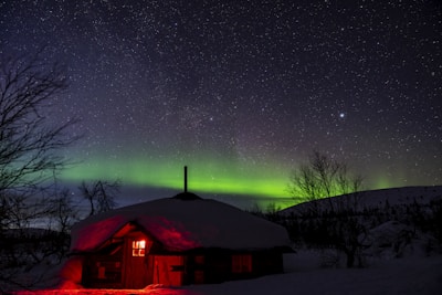 A snow-covered cabin softly illuminated by a red light sits amid trees under a vibrant night sky filled with stars. The aurora borealis casts a striking greenish-yellow glow across the horizon, complementing the dark silhouette of the cabin and surrounding landscape.