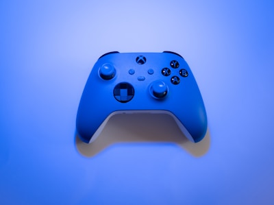 A video game controller with a sleek design and rounded edges, featuring buttons labeled X, Y, A, B, and a directional pad. The controller is illuminated by a soft blue light, giving it a futuristic appearance against the gradient blue background.