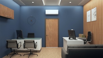A modern office interior with blue walls and wooden accents. The room features two desks, each with a computer and office chairs. A contemporary clock is mounted on the wall above one of the desks. The flooring is light wood, and framed art decorates the wooden paneled wall. There is a gray sofa, and an air conditioning unit is installed above a wooden door.