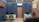 A modern office interior with blue walls and wooden accents. The room features two desks, each with a computer and office chairs. A contemporary clock is mounted on the wall above one of the desks. The flooring is light wood, and framed art decorates the wooden paneled wall. There is a gray sofa, and an air conditioning unit is installed above a wooden door.