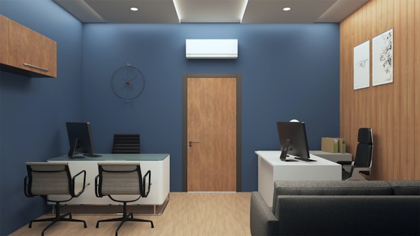 A modern office interior with blue walls and wooden accents. The room features two desks, each with a computer and office chairs. A contemporary clock is mounted on the wall above one of the desks. The flooring is light wood, and framed art decorates the wooden paneled wall. There is a gray sofa, and an air conditioning unit is installed above a wooden door.