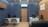 A modern office interior with blue walls and wooden accents. The room features two desks, each with a computer and office chairs. A contemporary clock is mounted on the wall above one of the desks. The flooring is light wood, and framed art decorates the wooden paneled wall. There is a gray sofa, and an air conditioning unit is installed above a wooden door.