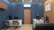 A modern office interior with blue walls and wooden accents. The room features two desks, each with a computer and office chairs. A contemporary clock is mounted on the wall above one of the desks. The flooring is light wood, and framed art decorates the wooden paneled wall. There is a gray sofa, and an air conditioning unit is installed above a wooden door.