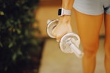 A person is holding a silver dumbbell with their right hand, wearing a watch with a light-colored band. The person is dressed in light blue shorts, and the background is a mix of greenery and a muted surface.