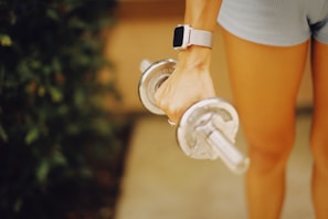 A person is holding a silver dumbbell with their right hand, wearing a watch with a light-colored band. The person is dressed in light blue shorts, and the background is a mix of greenery and a muted surface.
