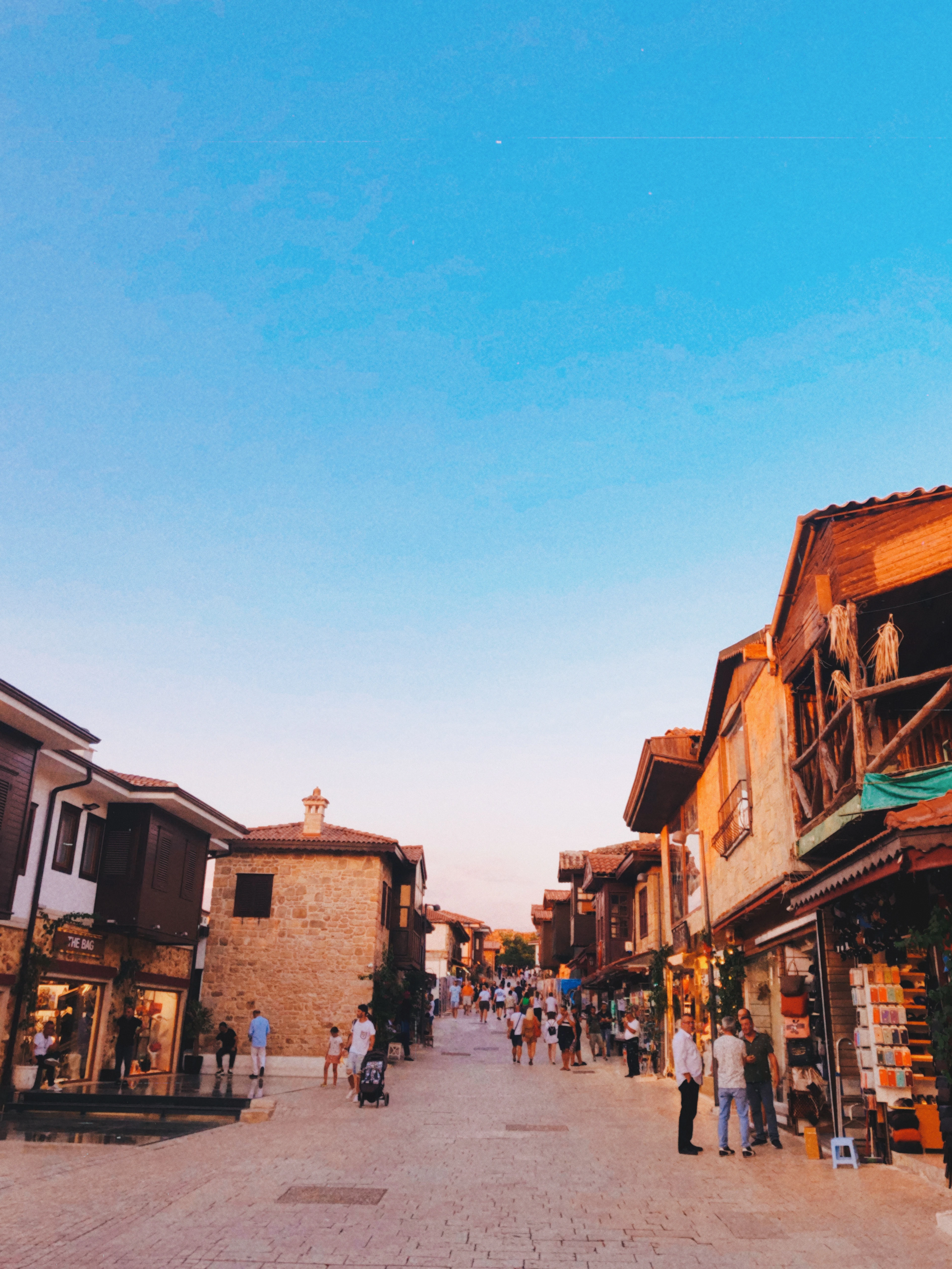 A cobbled Kaleiçi street lined with Ottoman-era houses and shops, golden evening light and strollers passing by.