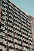 A modern, high-rise residential building with numerous balconies. The facade features a grid-like pattern with glass railings and dark panels. The sky in the background appears clear and blue.