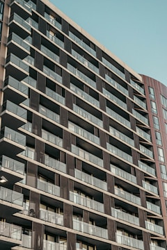 A modern, high-rise residential building with numerous balconies. The facade features a grid-like pattern with glass railings and dark panels. The sky in the background appears clear and blue.
