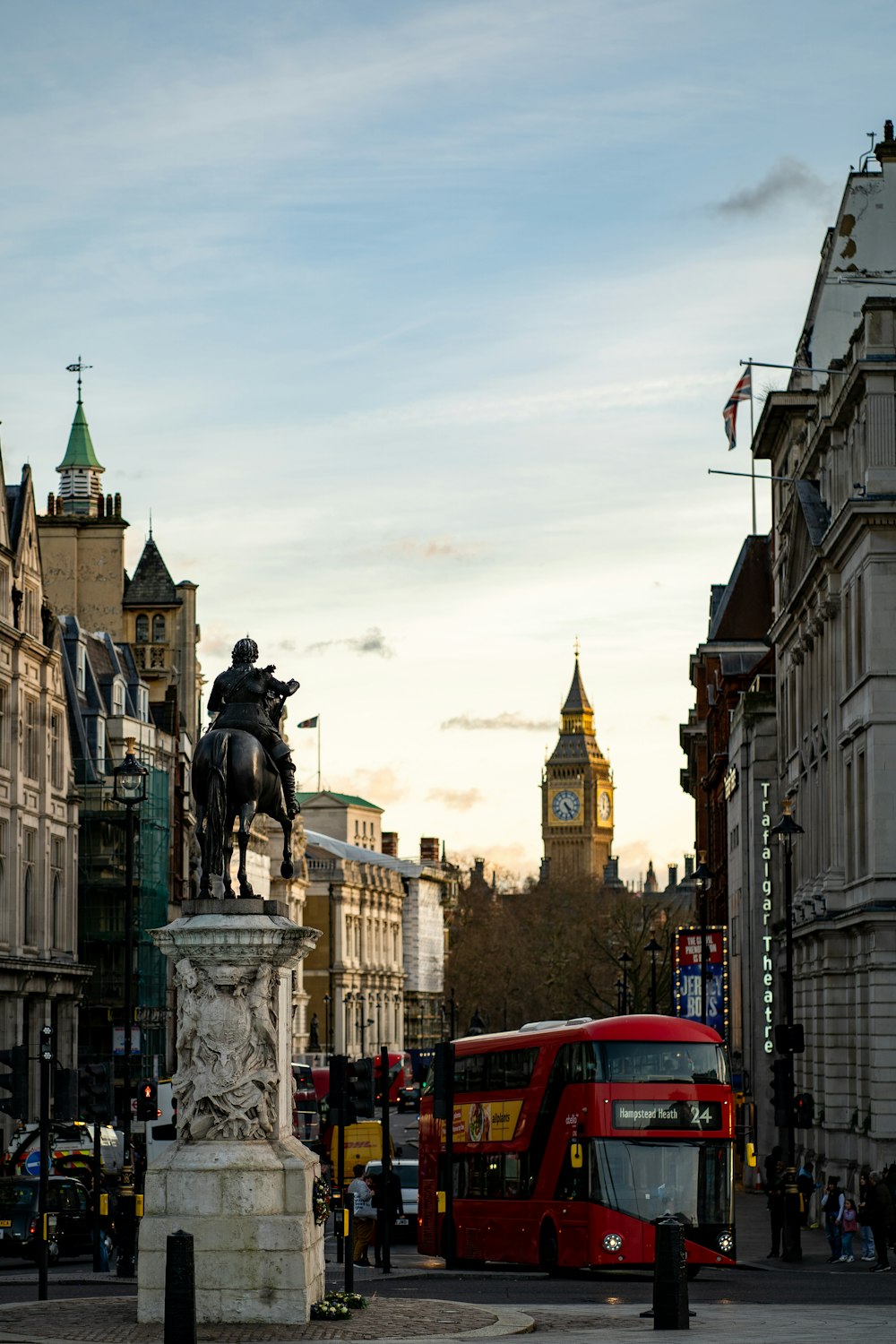a red double decker bus driving past a statue of a man on a horse