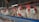 Several white chickens with red combs are standing inside a metal cage in a row. They are positioned side by side, showing limited space. The background includes a wooden structure of a typical poultry farm setup.