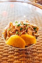 A small bowl filled with shredded meat, topped with finely chopped green onions and accompanied by two large slices of yellow pickled radish, placed on a woven, glass-topped table.