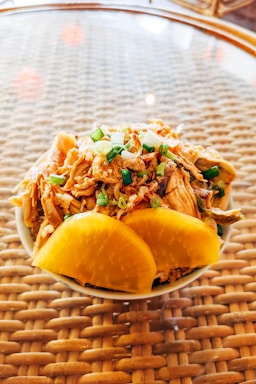 A small bowl filled with shredded meat, topped with finely chopped green onions and accompanied by two large slices of yellow pickled radish, placed on a woven, glass-topped table.