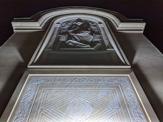 A stone relief sculpture with intricate carvings in a building façade. The relief depicts a religious or historical scene with figures in a deeply recessed niche. The architecture surrounding the sculpture includes geometric and decorative patterns, highlighted by soft lighting against a dark night sky.