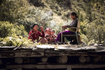 A family scene outdoors with an elderly person and children sitting on a wooden structure surrounded by lush greenery. The family appears engaged and content, with one person seated on a chair.
