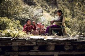 A family scene outdoors with an elderly person and children sitting on a wooden structure surrounded by lush greenery. The family appears engaged and content, with one person seated on a chair.