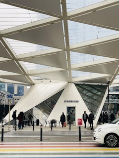 A modern architectural structure with a geometric design houses a Starbucks. People are seen walking and standing near the entrance, as well as some outdoor seating with tables and chairs. The setting appears to be urban with a crosswalk and street in the foreground.